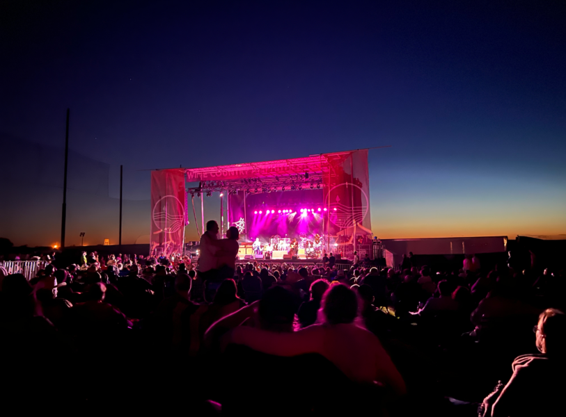 A wide shot of an outdoor concert crowd watching a band perform on a large stage lit with bright pink and purple lights against a deep blue sunset sky.