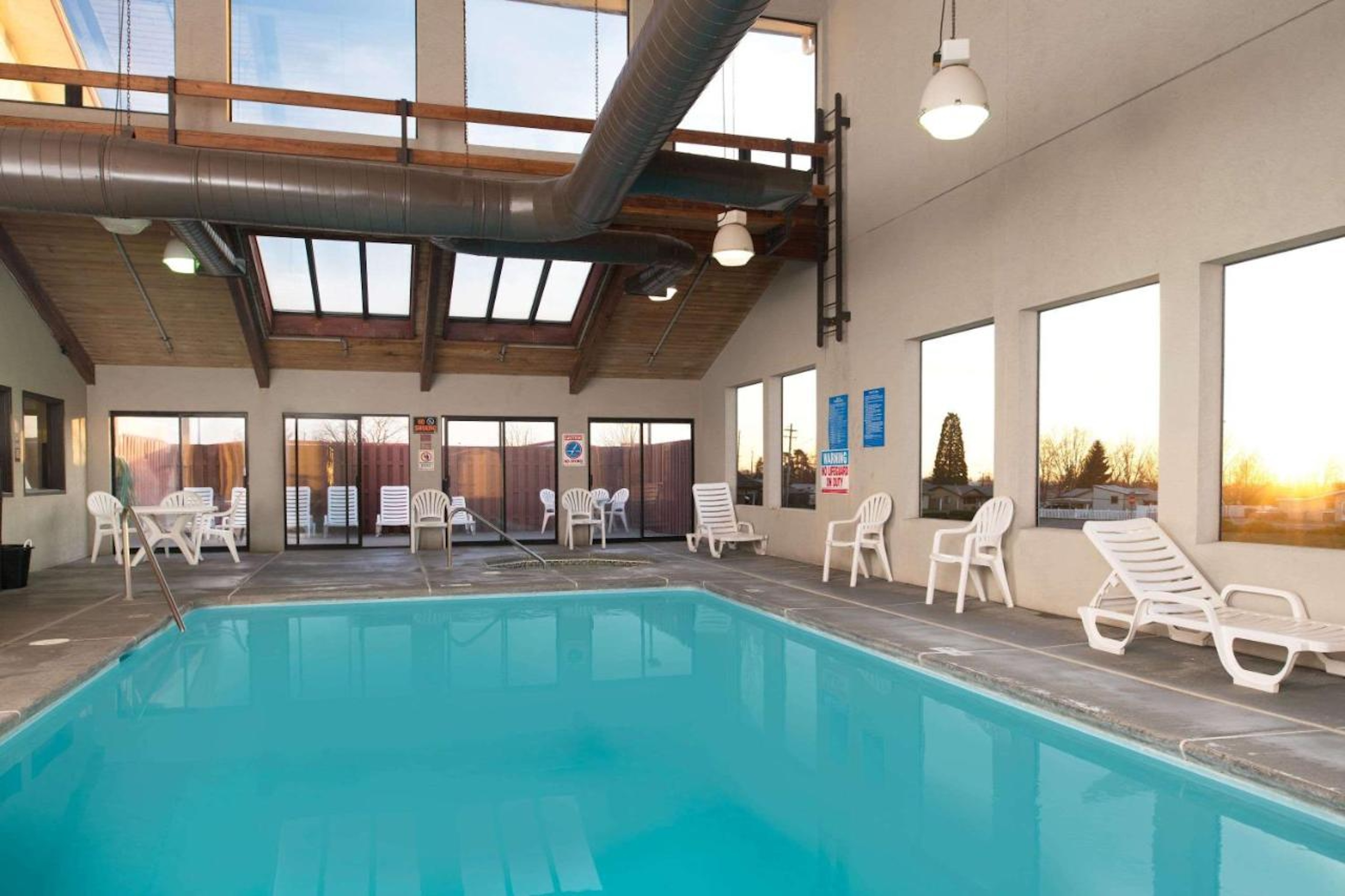 Indoor hotel pool with lounge chairs and large windows letting in sunset light.