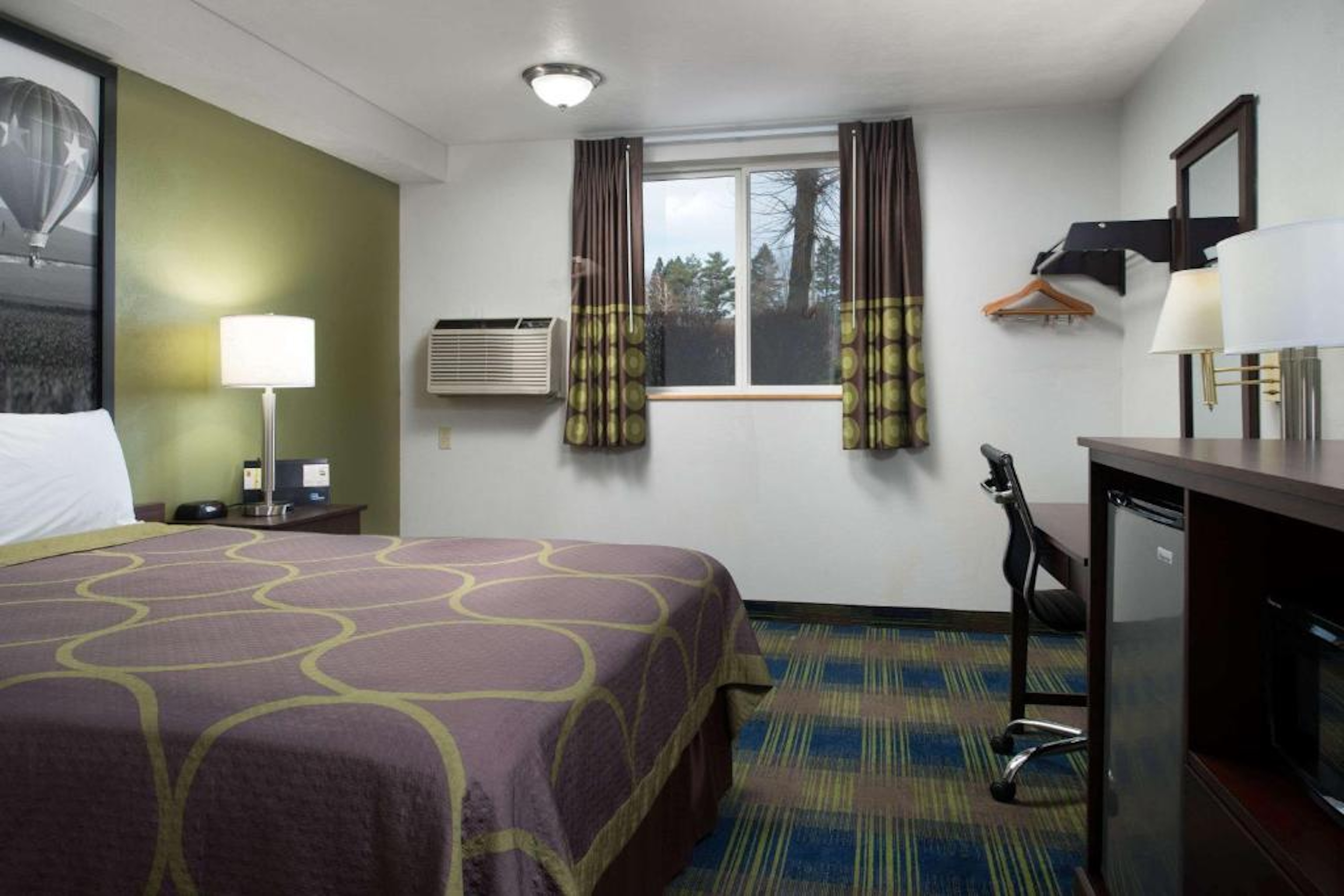 Hotel room with a queen bed, desk, mini fridge, and window with patterned curtains.