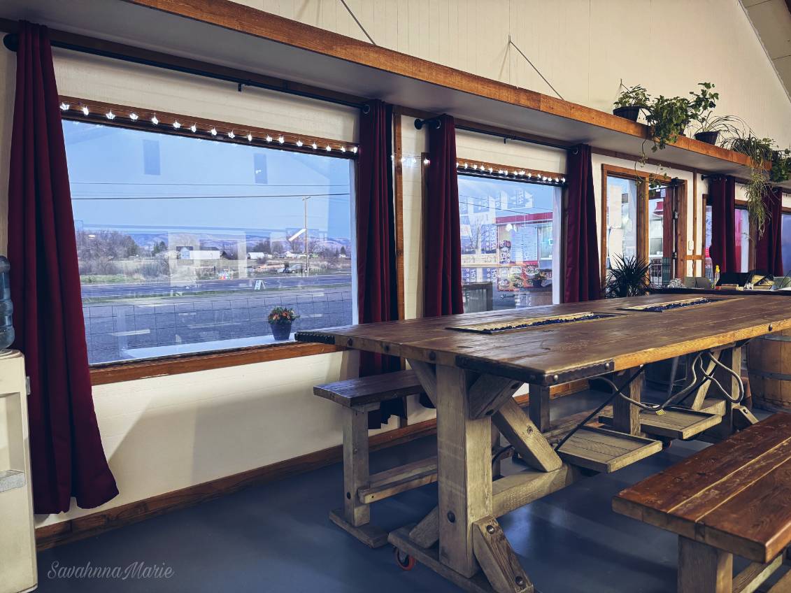 Rustic communal wooden table beside large windows with red curtains and potted plants.