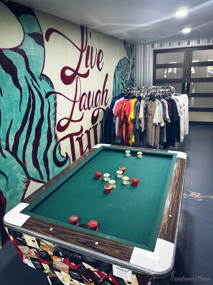 Shuffleboard table in front of a wall mural and clothing rack with t-shirts.