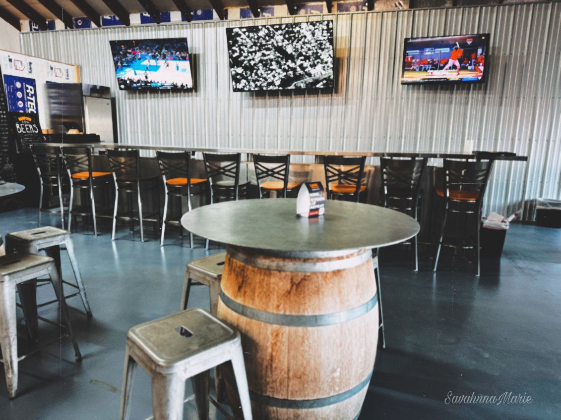 Sports bar with a long high-top bar, multiple TVs on a corrugated metal wall, and seating made from barrels and stools.