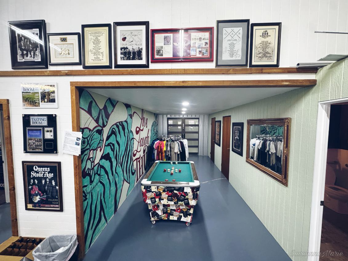 Pool table in a hallway with a “Live Laugh Thug” mural and hanging shirts.
