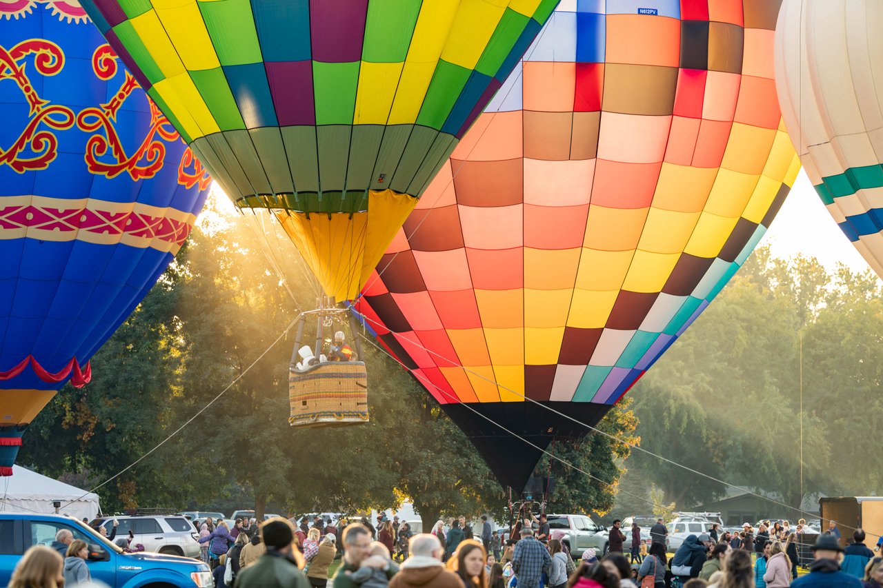 Colorful hot air balloons are being inflated over a crowd of spectators in a park at sunrise. Sunlight streams through the vibrant fabric.