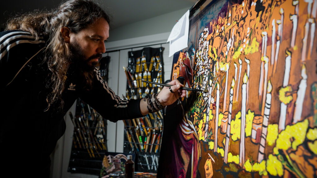 An artist with long hair and a beard paints abstract figures in vibrant orange and yellow hues onto a large, dark canvas in his studio.