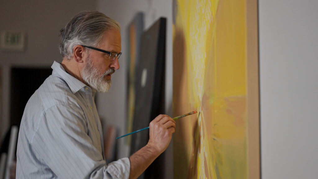 A distinguished, bearded man wearing glasses and a light shirt paints a large abstract canvas in shades of yellow and orange in his art studio.